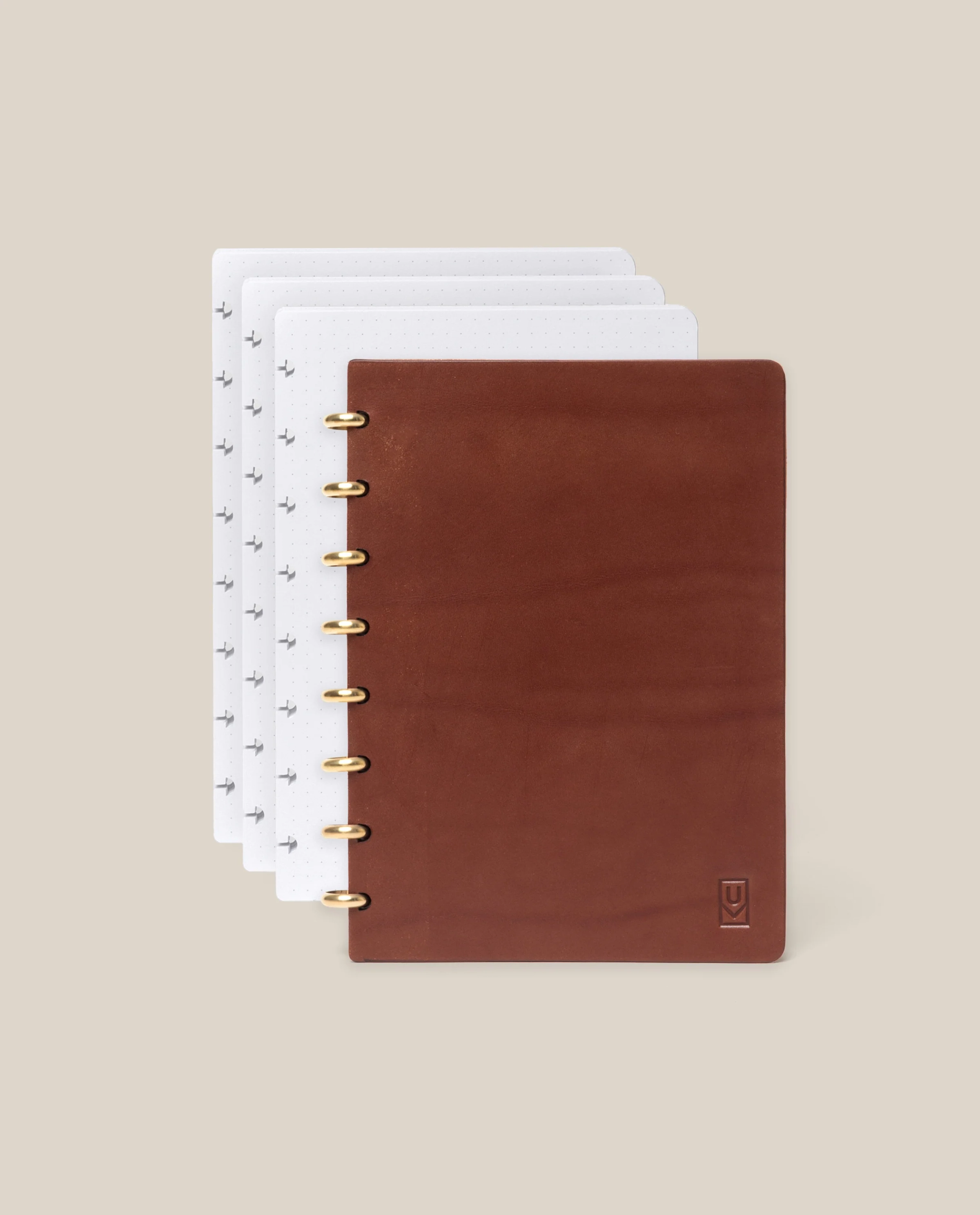 A Discbound Heirloom Journal + 3 Refills (Brown Leather 6″ x 8.5″), featuring full-grain brown leather and five gold discbinding rings, is partially open to display its dotted, symbol-printed white pages against a beige background.