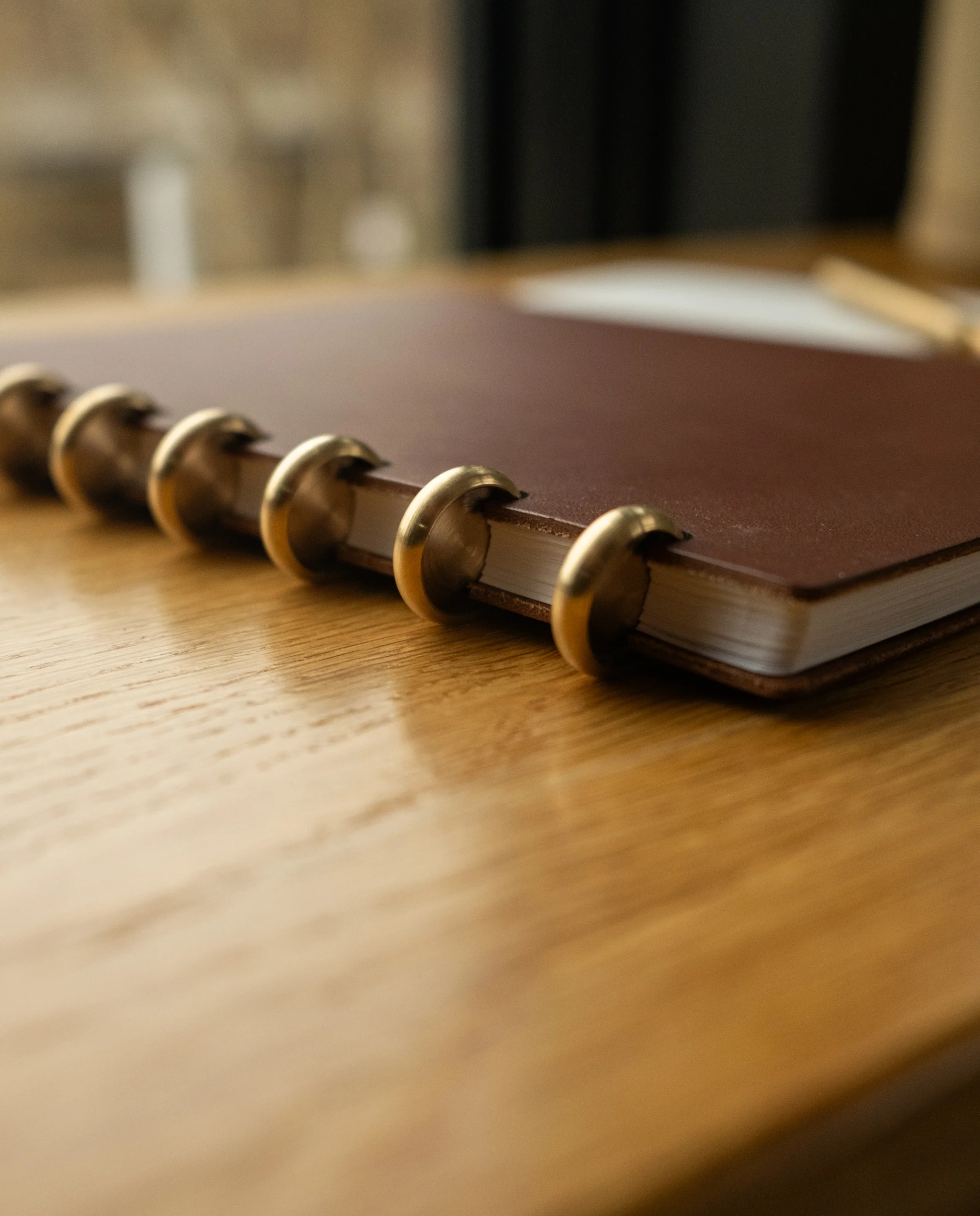 A Discbound Heirloom Journal + 3 Refills in brown leather (6″ x 8.5″) with large gold rings rests on a wooden desk, lying slightly open to reveal its pages, while the background remains gently out of focus.