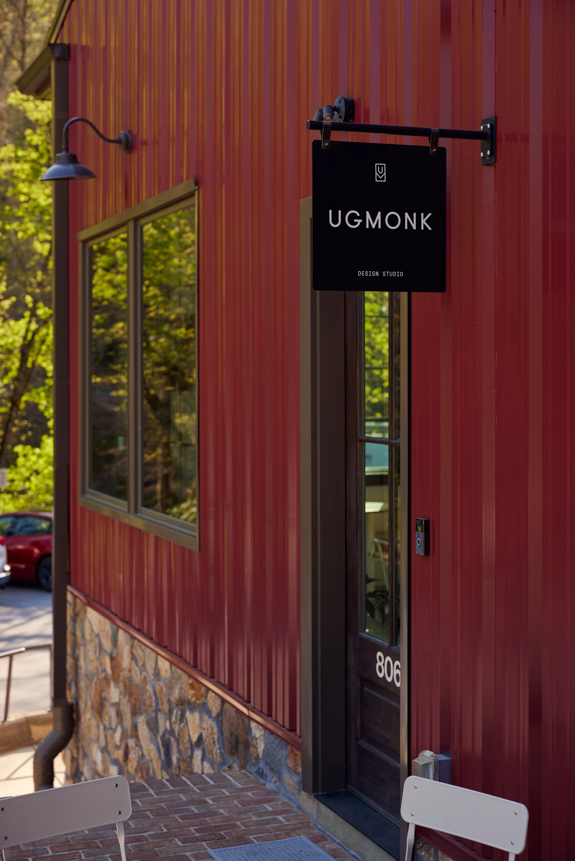 A red building with a stone base has a window and a door labeled 806. A black sign above the door reads UGMONK DESIGN STUDIO. Outdoor chairs are visible in the foreground.