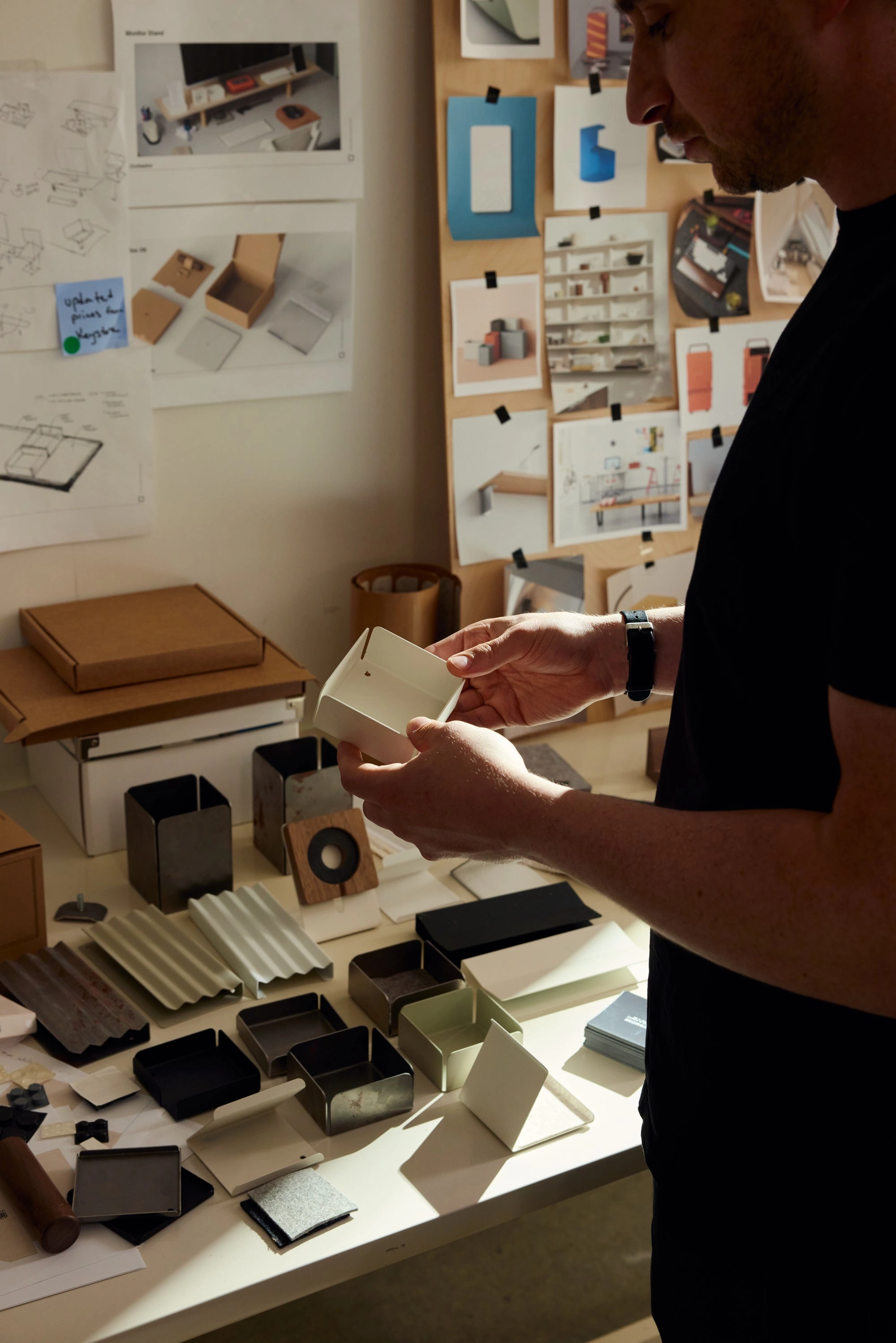 A person stands at a desk examining a small box, surrounded by various packaging prototypes, materials, and design sketches pinned to the wall.