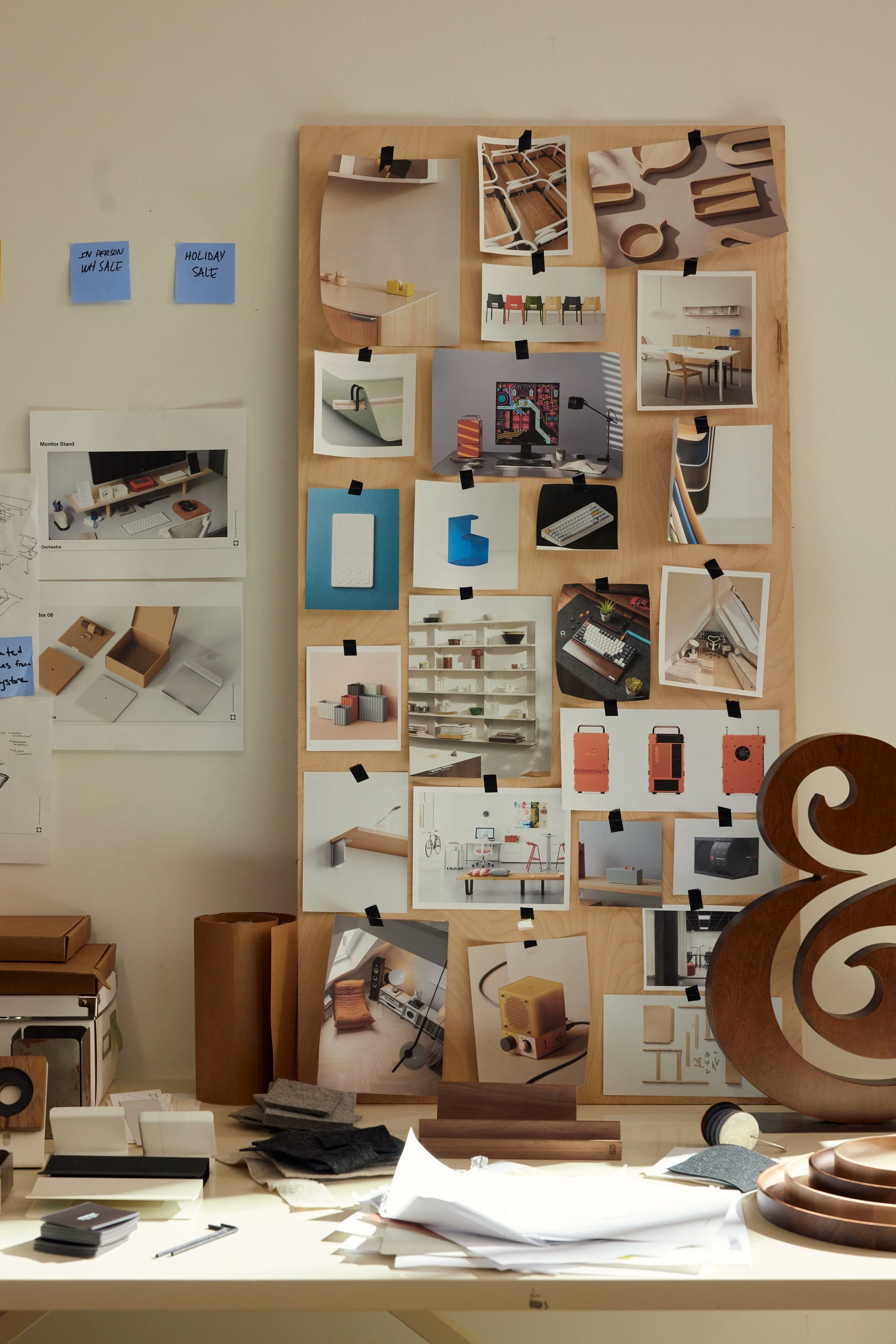 A mood board with various product and interior photos is propped on a cluttered desk with papers, samples, and a wooden ampersand decoration. Blue sticky notes and sketches are pinned to the wall beside it.
