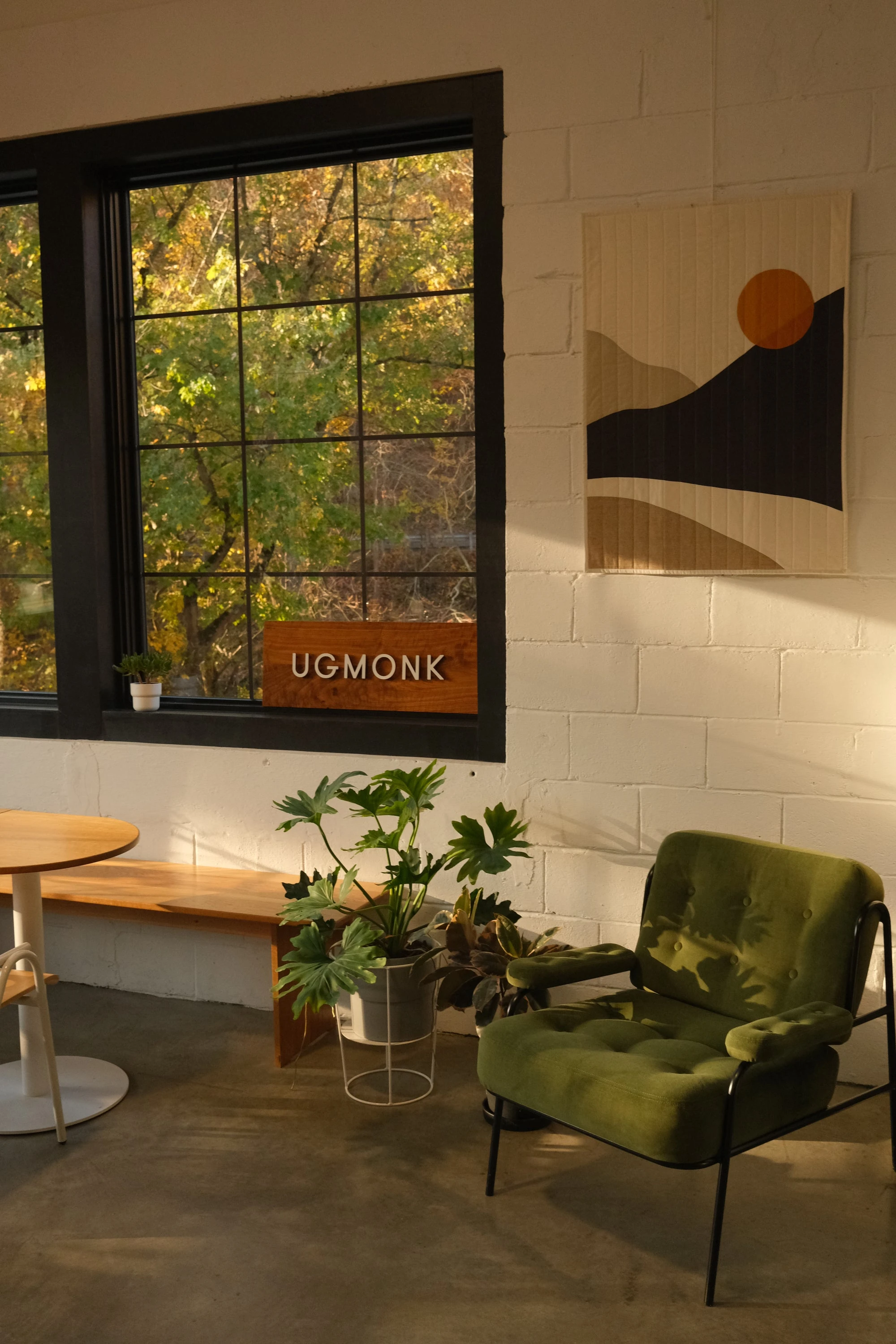 Sunlit modern room with a green armchair, small round table, plants, and wall art. Large windows reveal trees with green leaves outside. A wooden sign reading UGMONK sits on the bench below the window.